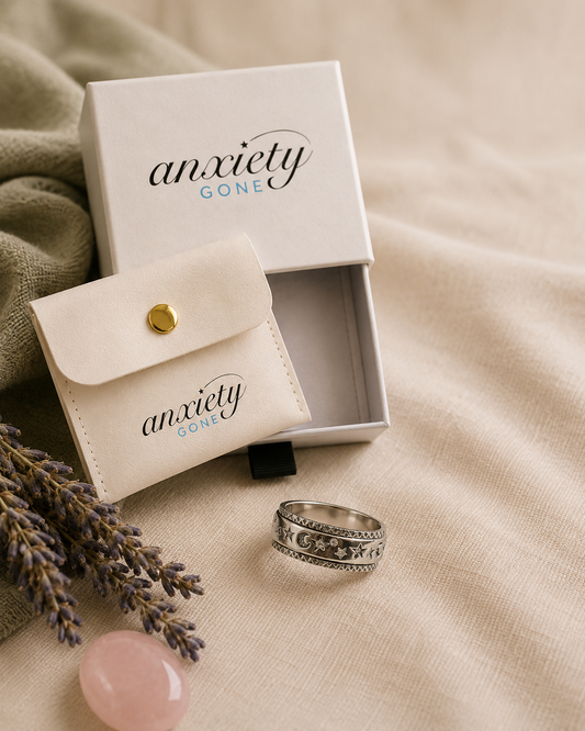 An ornate silver fidget ring with engraved patterns is displayed next to its cream-colored velvet pouch and matching white presentation box, both printed with the brand name 'anxiety GONE'. The scene is styled on a beige fabric background with lavender sprigs and a pink stone.