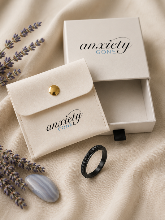 A cream-colored jewelry box and matching card case with the brand name 'anxiety GONE' printed on them, styled on a beige fabric background with lavender sprigs and a black fidget ring.