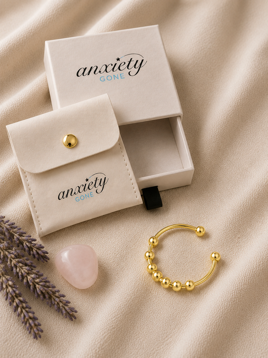 A gold beaded open-ended spinner ring displayed next to its cream-colored branded storage pouch and box on a beige fabric background, with decorative lavender sprigs and a pink stone.