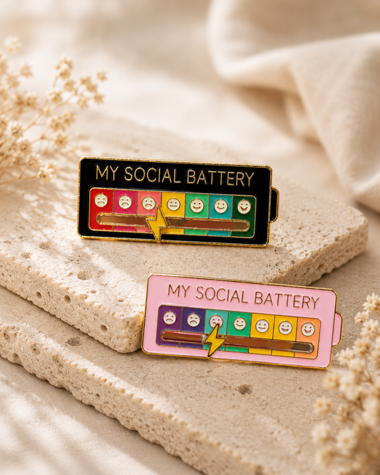 Two enamel battery-shaped pins with the text 'MY SOCIAL BATTERY' rest on a textured stone slab with soft beige fabric in the background. One pin is black with a yellow battery level and mostly empty smiley faces, and the other is pink with a yellow battery level and several sad smiley faces.