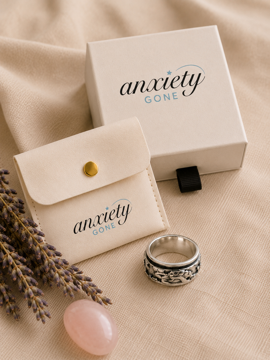 Anxiety Gone brand jewelry packaging set with a cream velvet pouch, a cream gift box, and a silver patterned turtle fidget ring on a beige fabric background with lavender sprigs and a pink stone.