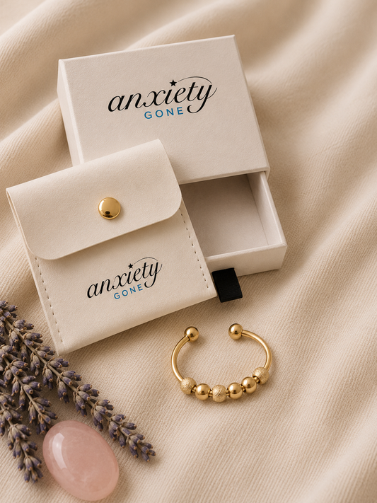 A gold beaded fidget ring displayed next to its cream-colored branded storage pouch and matching box on a beige fabric background, with lavender sprigs and a pink stone as props.