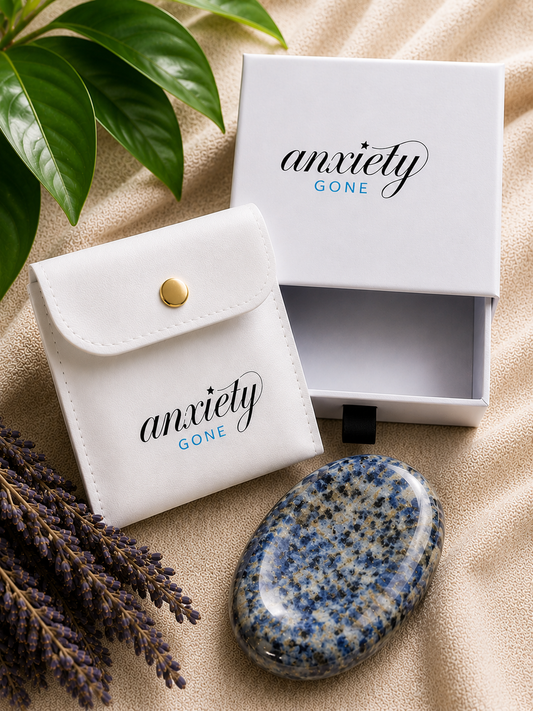 Anxiety Gone branded white gift set including a small white pouch with a gold snap, an open white box, and a polished blue-and-white speckled stone, styled on beige fabric with green leaves and lavender sprigs.