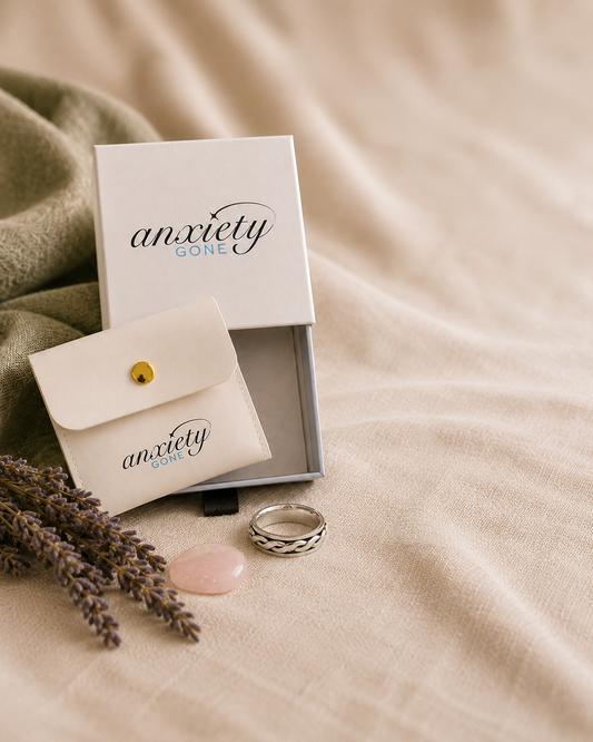 A silver patterned fidget ring displayed next to its cream-colored 'Anxiety Gone' gift box and matching pouch on a beige fabric background.