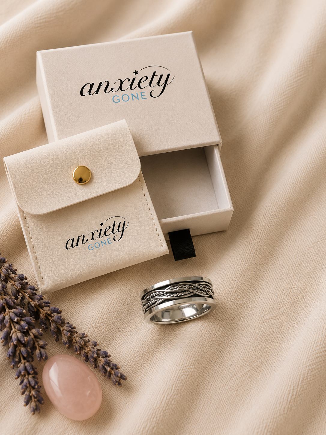 A silver fidget ring with a textured, wavy patterned band is displayed next to its cream-colored packaging, including a branded jewelry box and a matching pouch, on a beige fabric background with decorative lavender and a pink stone.