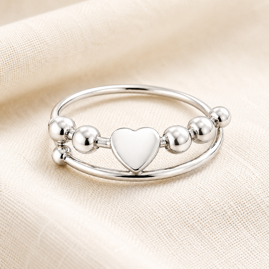 A polished silver-tone double-band heart fidget ring featuring a central heart charm and small metallic bead accents, displayed on a soft cream-colored textured fabric background.