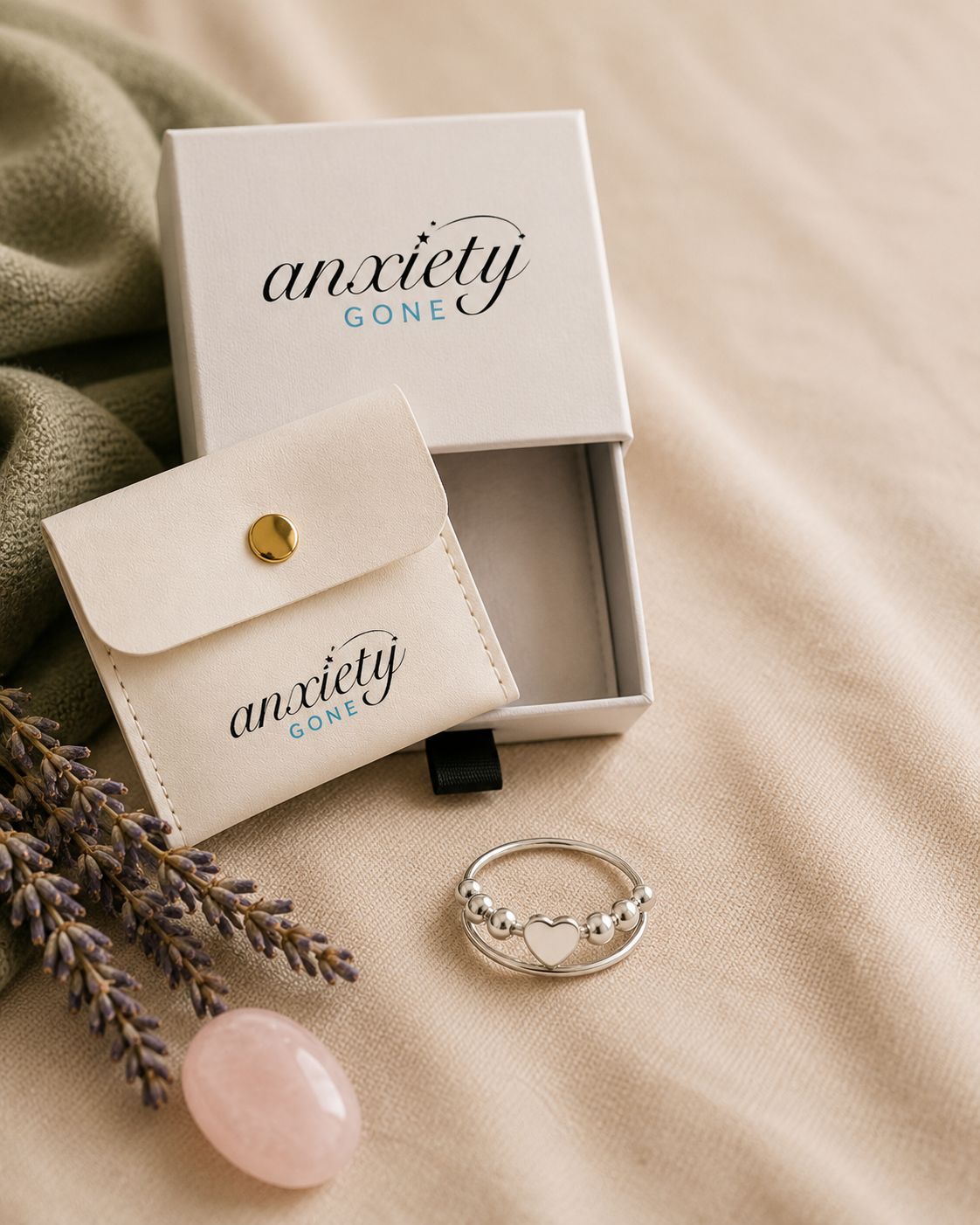 A silver heart fidget ring with a small heart and bead details is displayed next to its white 'anxiety GONE' branded gift box and pouch on a beige fabric background with decorative lavender and a pink stone.