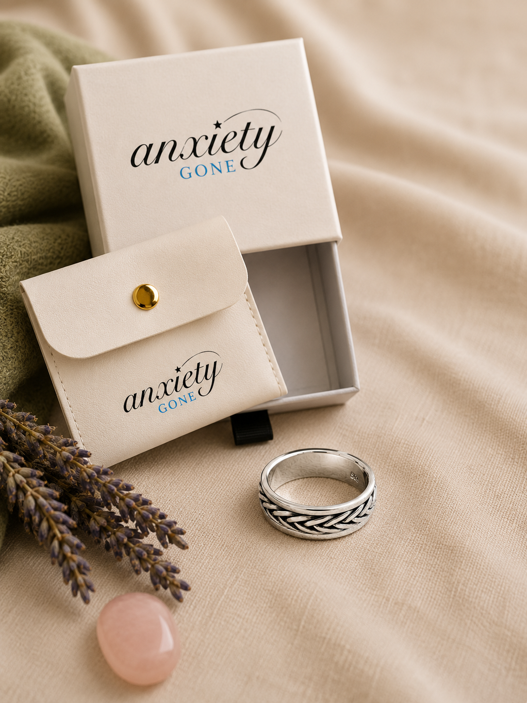 A silver fidget ring with an engraved geometric pattern, displayed next to its cream-colored branded 'anxiety GONE' gift box and pouch on a beige fabric background.