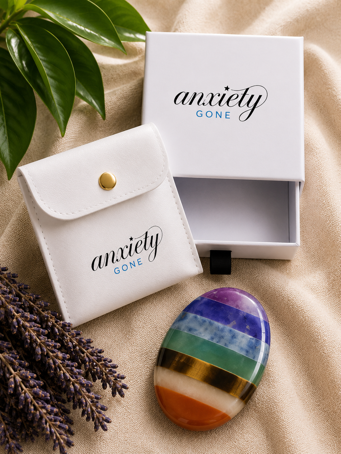 An oval rainbow chakra stone with polished bands of purple, blue, green, yellow, gold, and orange, displayed next to its white branded storage pouch and white gift box on a beige fabric background with decorative green leaves and lavender.