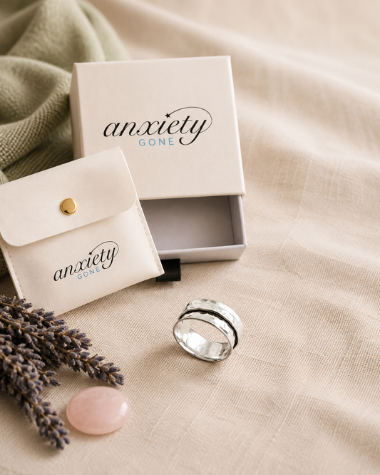 A silver spining band fidget ring with black inlay details, displayed next to its cream-colored 'Anxiety Gone' gift box and matching card on a beige linen fabric background.