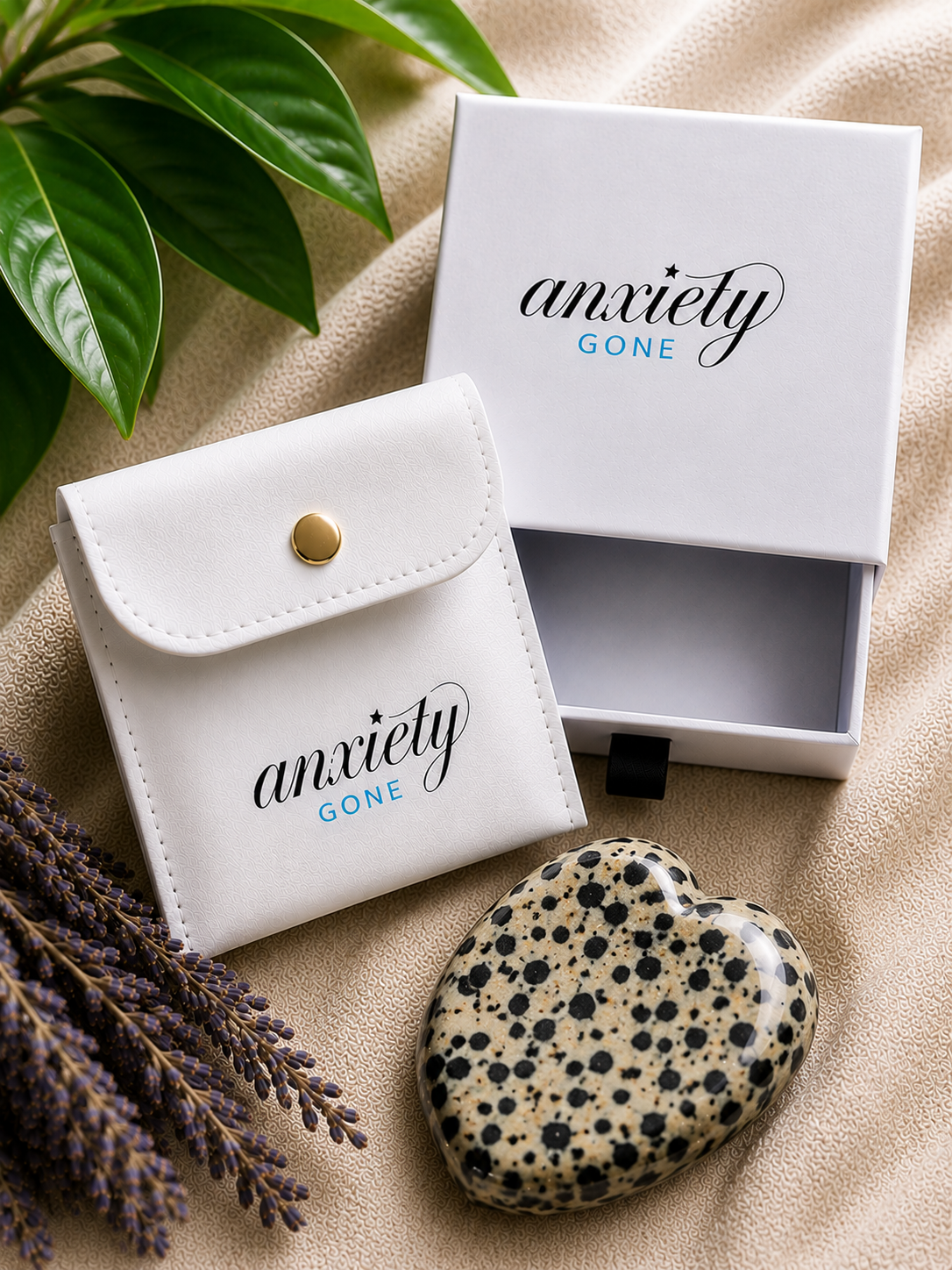 Anxiety Gone branded white pouch and matching box displayed on beige fabric with green leaves and lavender sprigs, alongside a speckled beige and black  dalmation jasper stone.