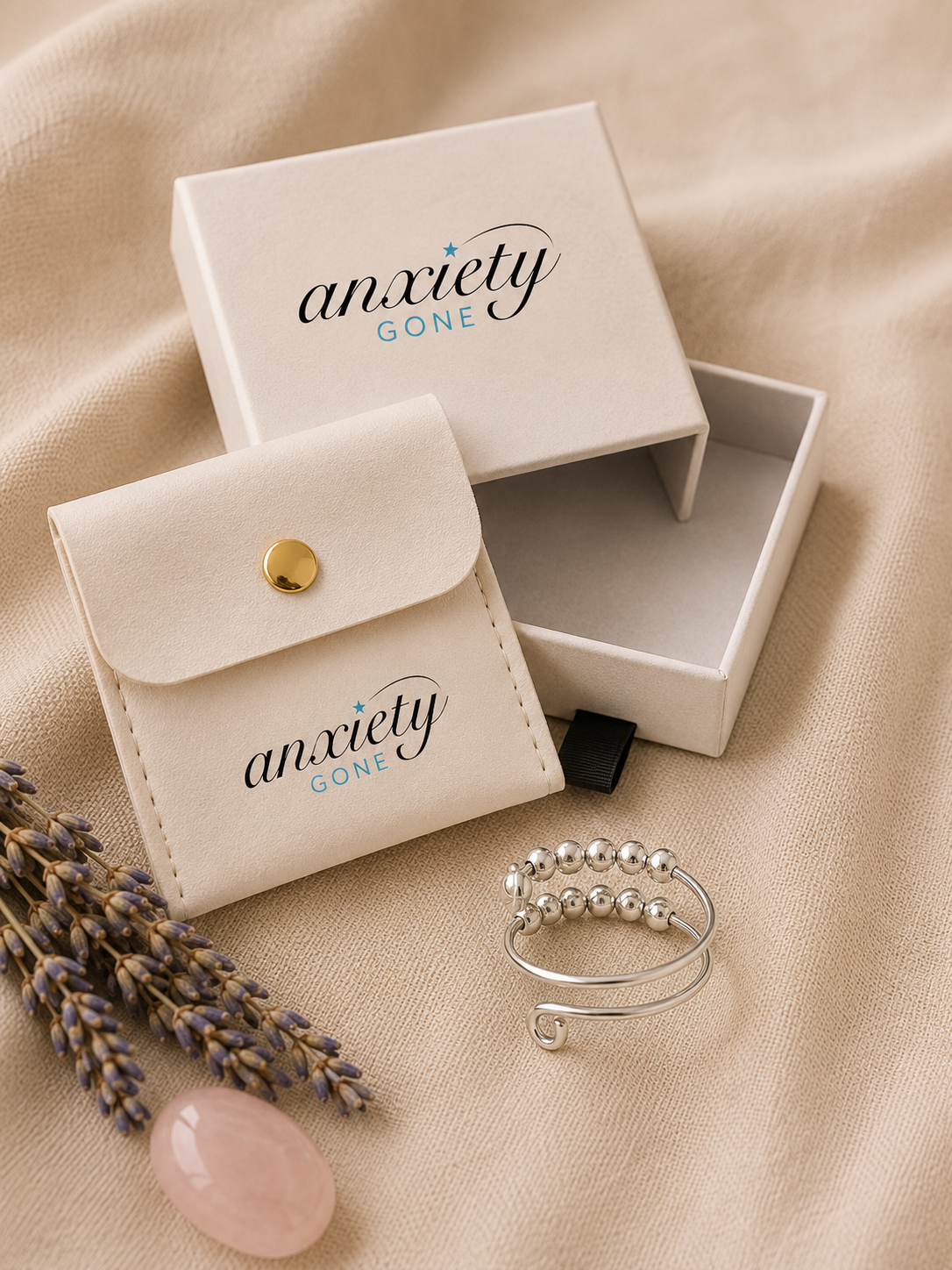 Anxiety Gone jewelry packaging set with a cream-colored box, matching pouch, and a silver beaded fidget ring on a beige fabric background with lavender sprigs and a pink stone.