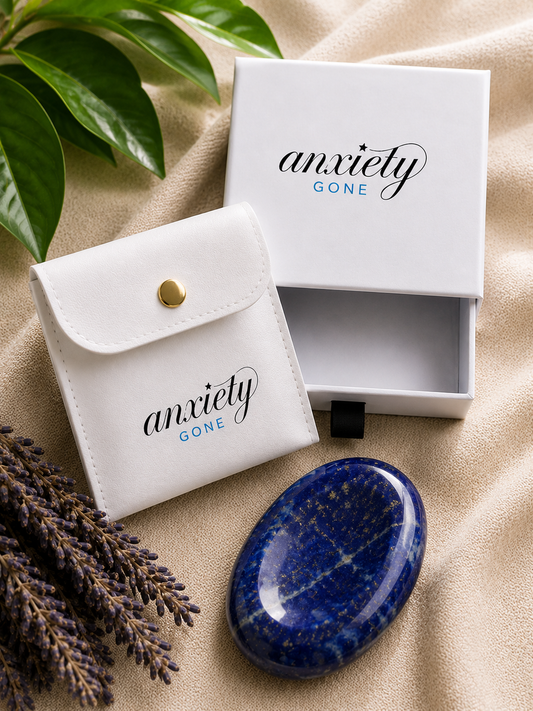 A polished blue selenite crystal with a star map pattern, displayed next to its white storage pouch and open presentation box, both featuring the 'anxiety GONE' logo, on a beige fabric background with decorative green leaves and lavender sprigs.