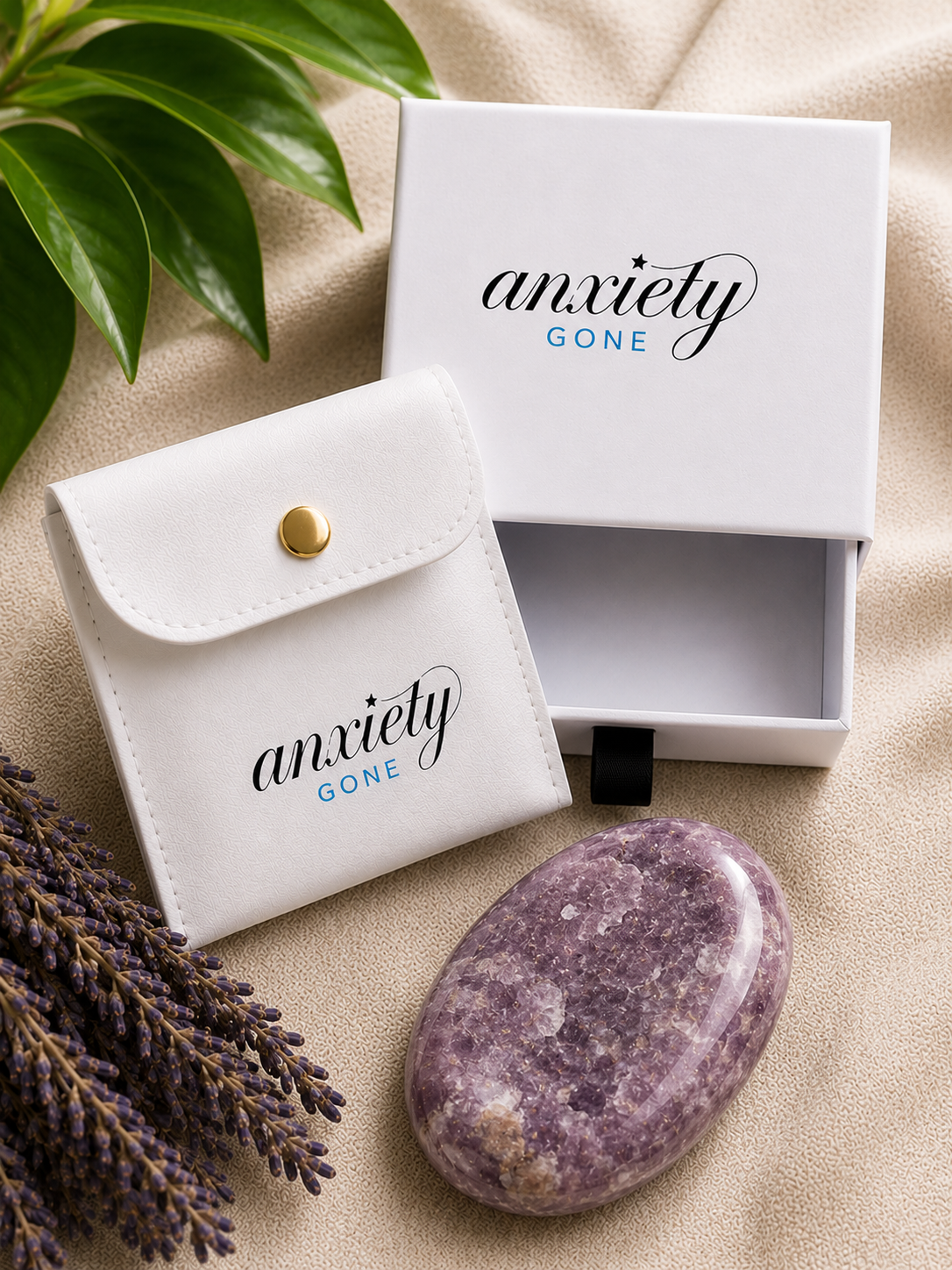 Anxiety Gone branded white gift set including a white pouch with a gold snap, a white box with the brand name, and a polished purple stone, styled on beige fabric with green leaves and lavender sprigs.