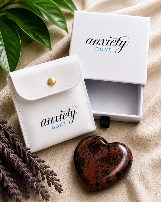 A polished brown and black speckled mahogany obsidian heart-shaped gemstone sits in front of a white branded pouch and matching open box labeled 'anxiety GONE' on a beige fabric surface with green leaves and lavender sprigs.