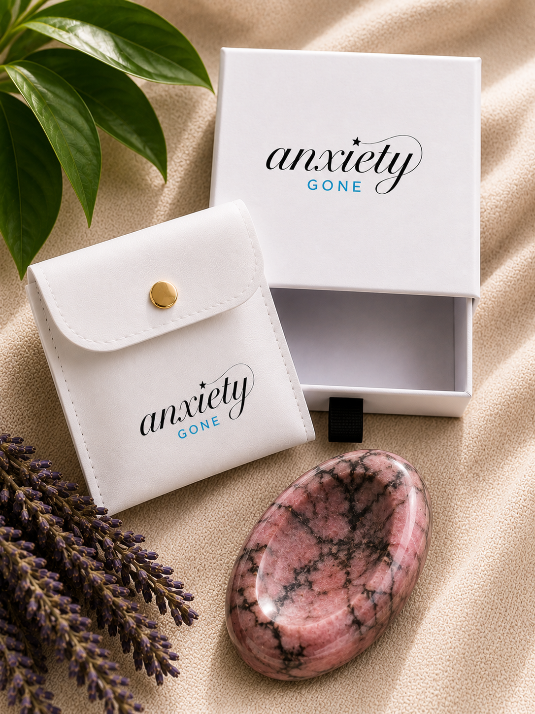 An oval pink and black marbled stone, likely a rose quartz crystal, displayed next to its white branded 'anxiety GONE' pouch and open presentation box on a beige fabric background with decorative green leaves and lavender.