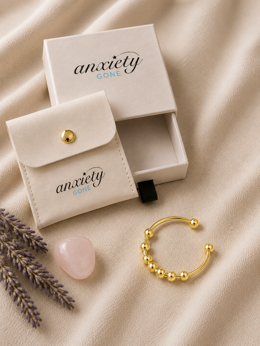 A gold beaded open-ended spinner ring displayed next to its cream-colored branded storage pouch and box on a beige fabric background, with decorative lavender sprigs and a pink stone.