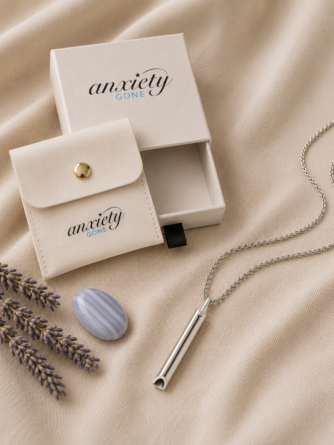 A silver breathing necklace on a chain, displayed with its cream-colored 'anxiety GONE' branded jewelry box and matching pouch on a beige fabric background.
