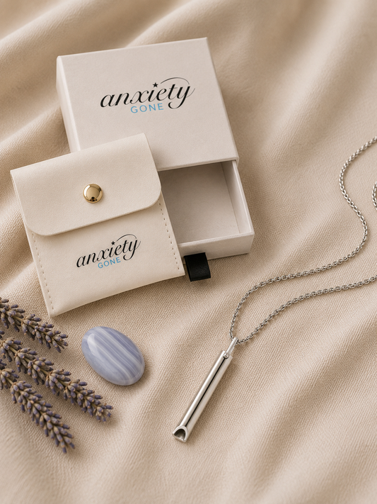 A silver breathing necklace on a chain, displayed with its cream-colored 'anxiety GONE' branded jewelry box and matching pouch on a beige fabric background.