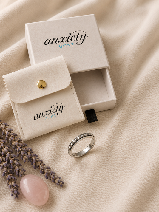A silver fidget ring with a pavé-set clear stone is displayed next to its cream-colored 'Anxiety Gone' branded jewelry box and matching pouch on a beige fabric background with lavender sprigs and a pink stone.