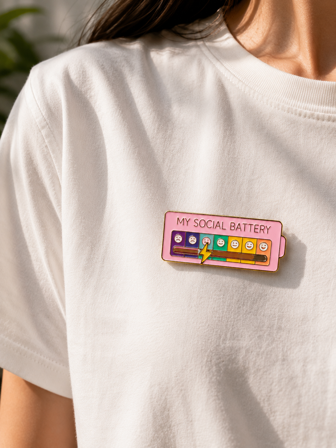 A close-up of a person wearing a white t-shirt with a colorful enamel pin shaped like a battery labeled 'MY SOCIAL BATTERY' on the chest.