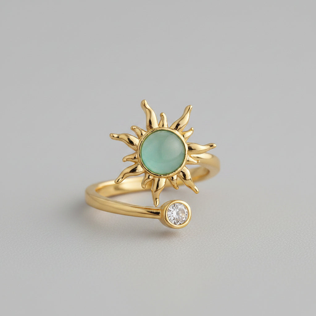 A gold fidget ring featuring a stylized sun design with a round light green stone in the center and a small clear gemstone on the band, set against a neutral gray background.