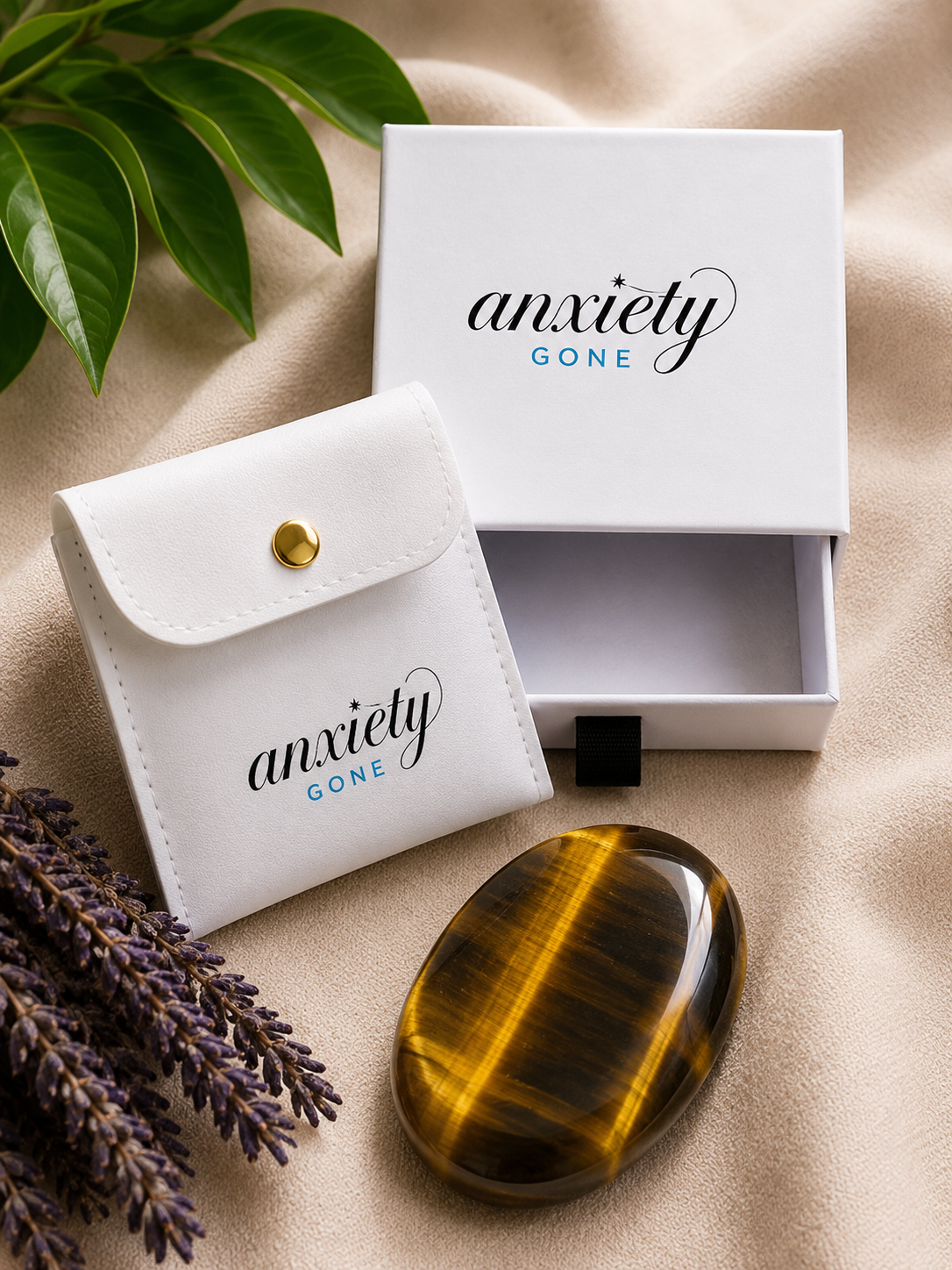 A polished tiger's eye stone with yellow and black bands, displayed next to its white branded 'anxiety GONE' gift box and matching white pouch on a beige fabric surface with green leaves and lavender sprigs.