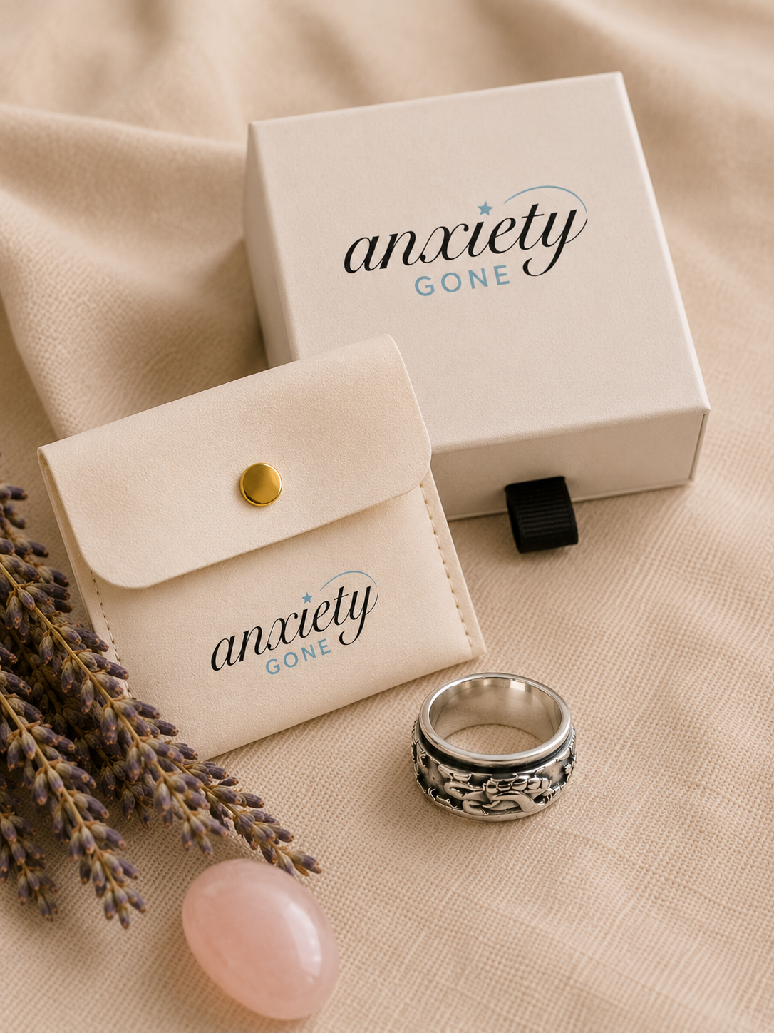 Anxiety Gone brand jewelry packaging set with a cream velvet pouch, a cream gift box, and a silver patterned turtle fidget ring on a beige fabric background with lavender sprigs and a pink stone.