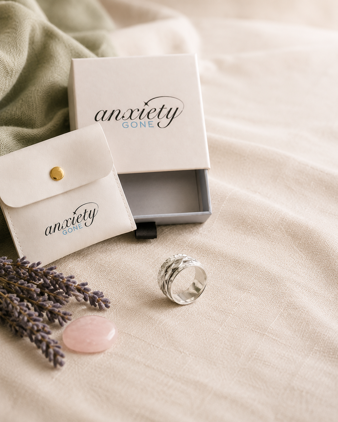 A silver textured fidget ring displayed next to its white 'Anxiety Gone' branded gift box and matching pouch on a beige fabric surface with lavender and a pink stone.