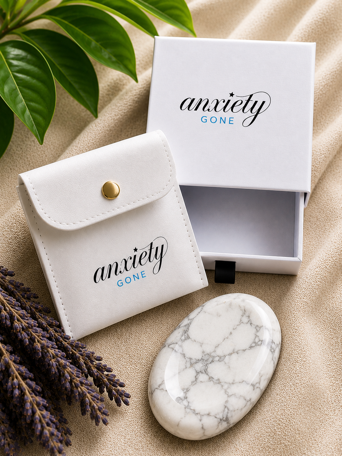 Anxiety Gone branded white gift set including a white storage pouch with a gold snap button, an open white presentation box, and a white howlite stress stone, styled on a beige fabric background with green leaves and lavender sprigs.