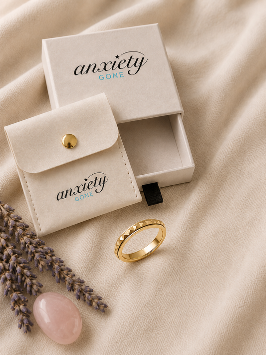 A gold fidget ring with a textured, beaded outer band is displayed next to its cream-colored presentation box and a matching velvet pouch on a beige fabric background, styled with lavender sprigs and a pink stone.